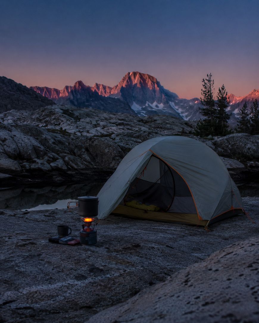 A tent is set up on rocky ground near a small lake, with a camp stove and mugs in front. Snow-capped mountains glow in the pink light of sunset in the background. Pine trees stand to the right.