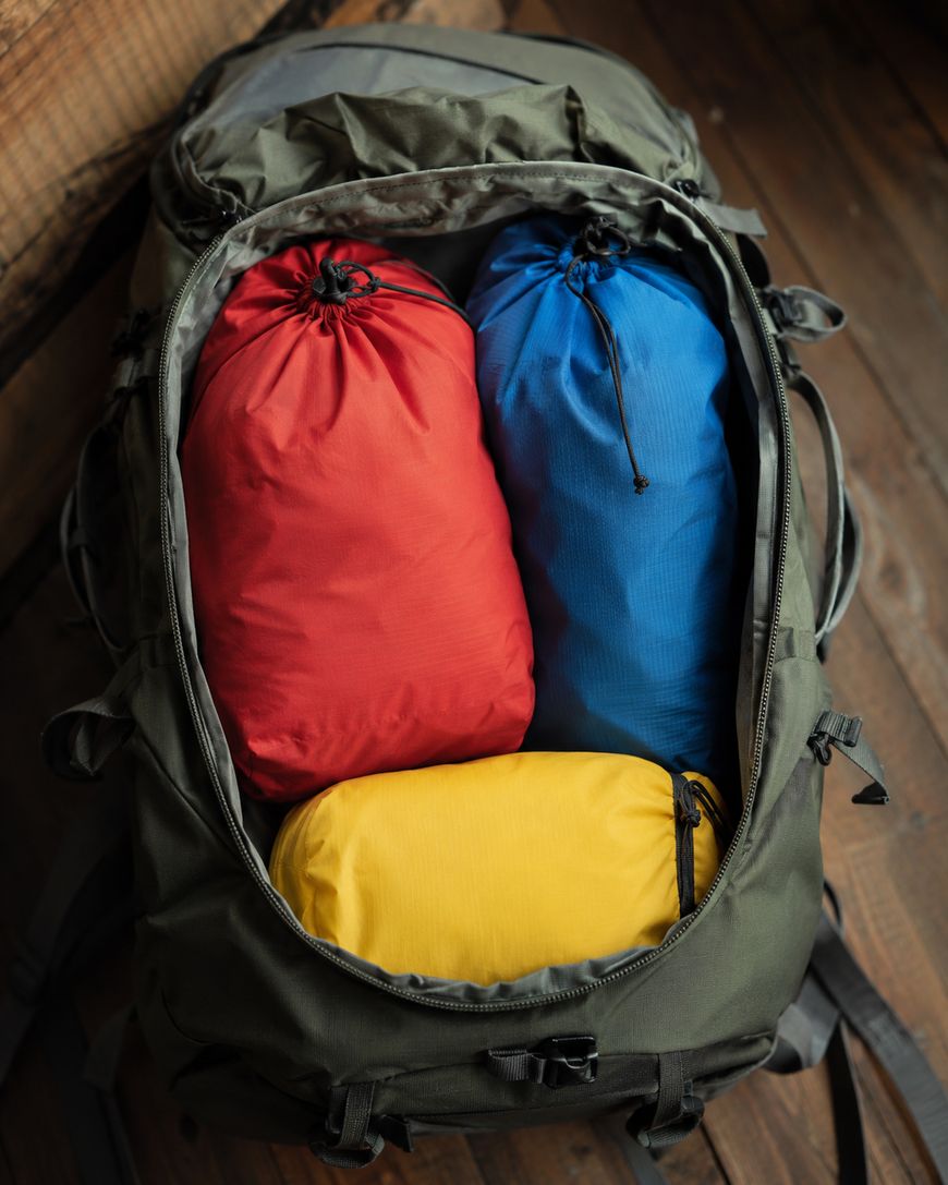 An open green backpack on a wooden surface, containing three packed bags: one red, one blue, and one yellow, neatly arranged inside.