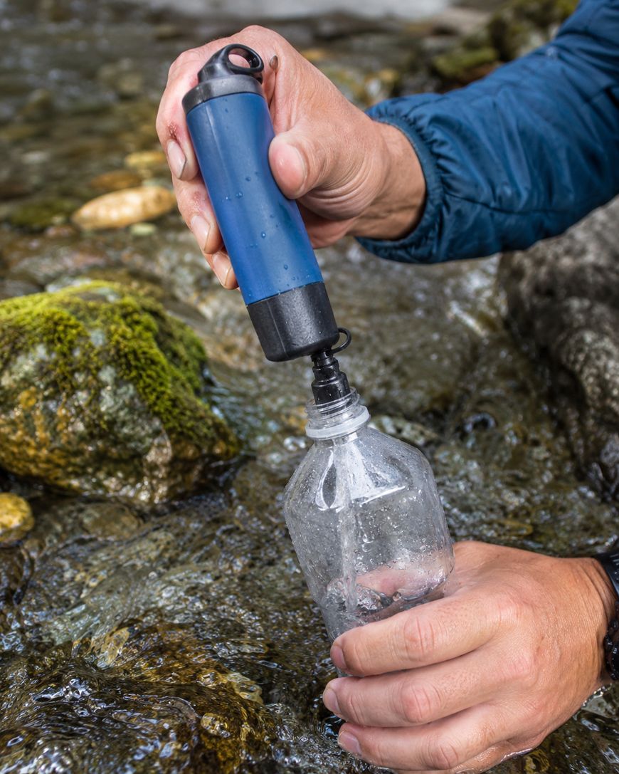 A person fills a plastic bottle with water from a stream using a blue portable water filter. Moss-covered rocks are visible in the flowing water.