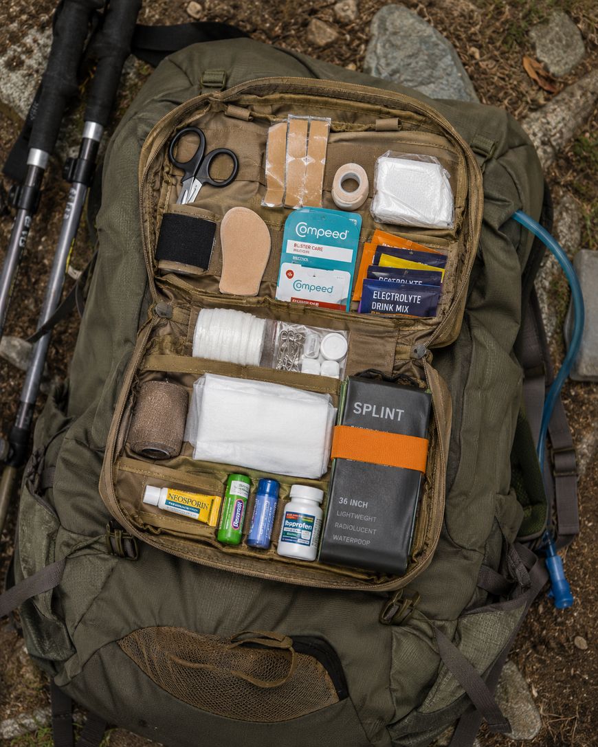 An open green backpack on the ground shows a neatly organized first aid kit with bandages, tape, scissors, gauze, medications, a splint, electrolyte drink mix, and other medical supplies inside a mesh compartment.