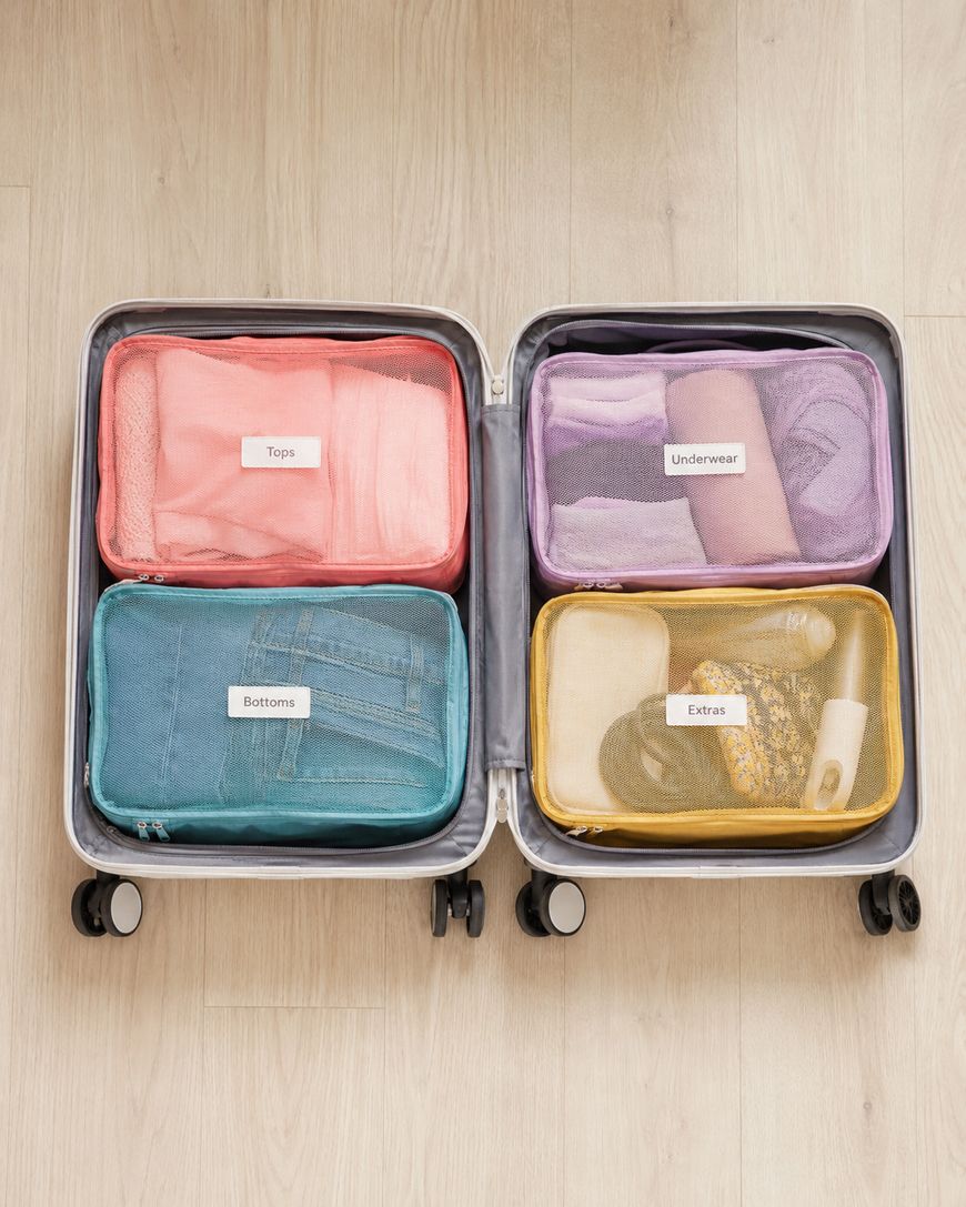 An open suitcase with four packing cubes labeled Tops, Underwear, Bottoms, and Extras, each containing neatly folded clothes and items, on a light wooden floor.