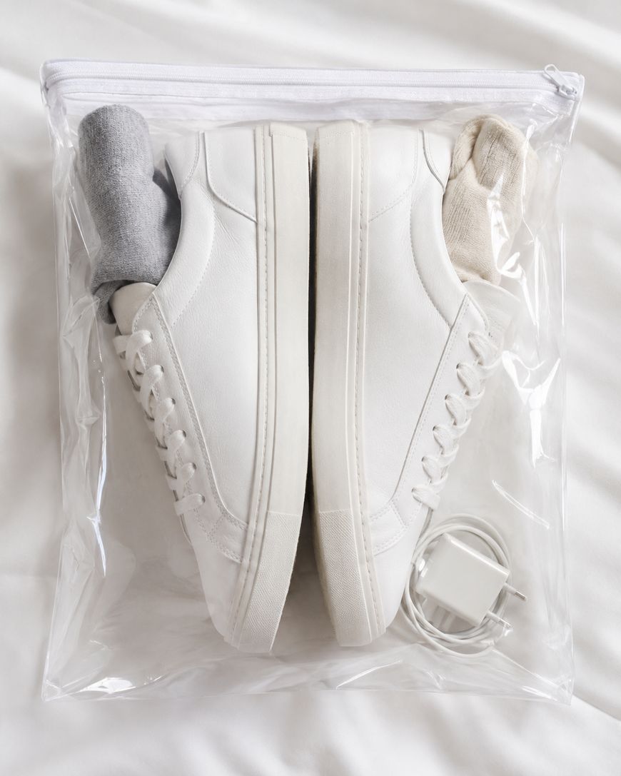 A pair of white sneakers with socks tucked inside, placed in a clear zippered plastic bag alongside a white charging cable, all on a white surface.