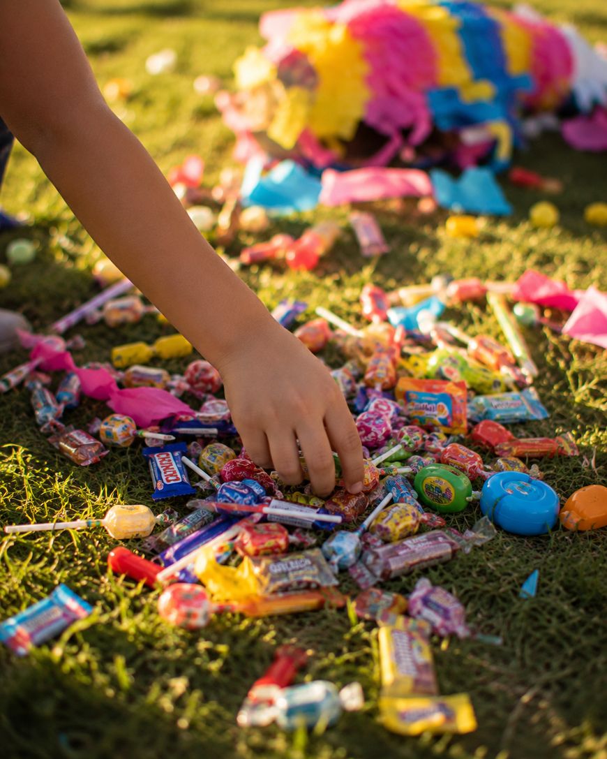 A child’s hand reaches for candies scattered on grass, spilled from a broken piñata in the background on a sunny day.