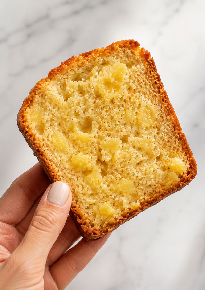A hand holds a slice of golden, moist pineapple bread with visible chunks of pineapple against a white marble background.