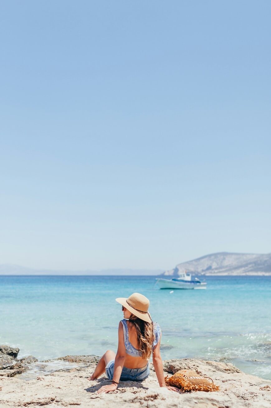A woman wearing a sunhat sits on a rocky shore, facing the turquoise sea with a boat in the distance and mountains under a clear blue sky. A woven bag rests beside her.