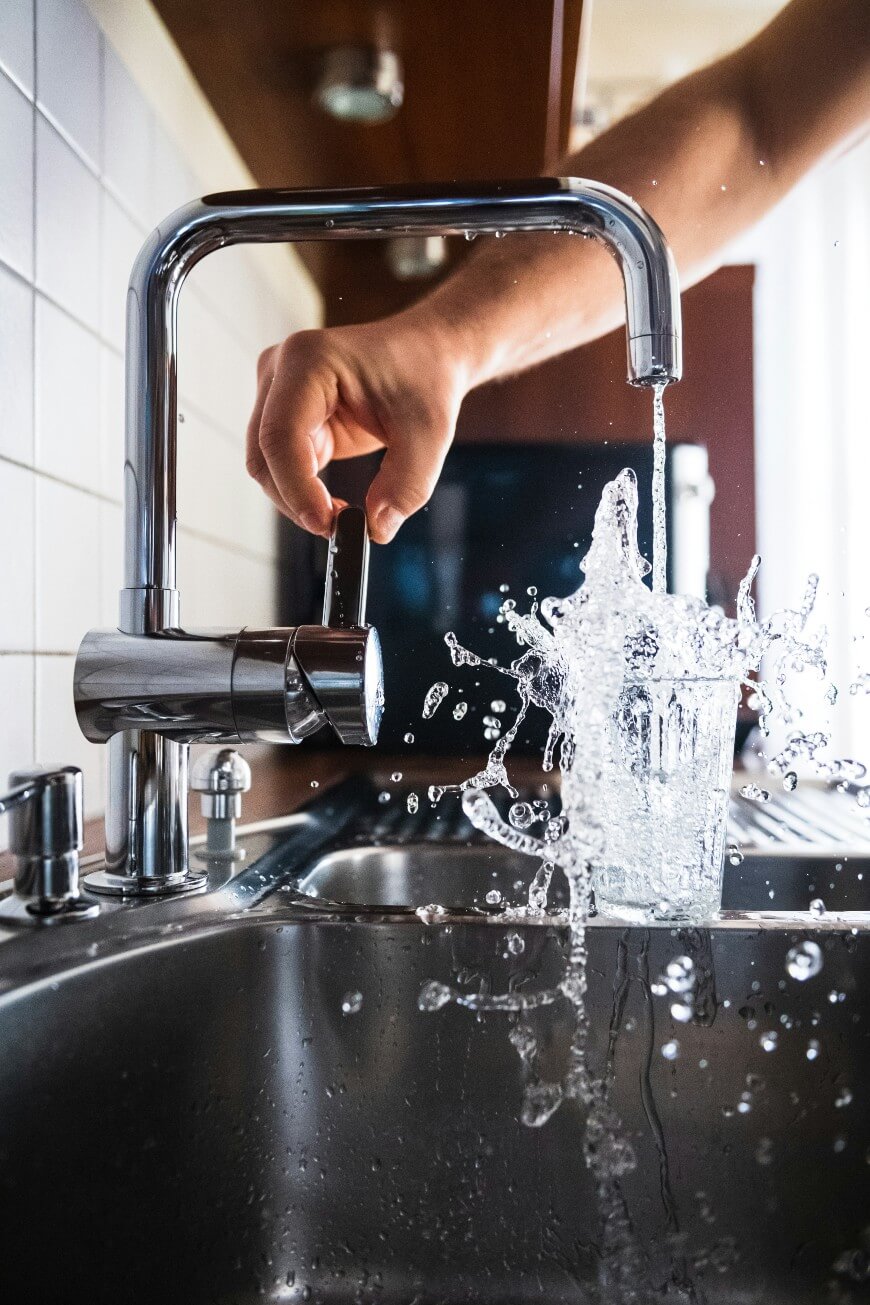 A hand adjusts a kitchen faucet as water splashes energetically into a glass, with droplets scattering over a stainless steel sink.