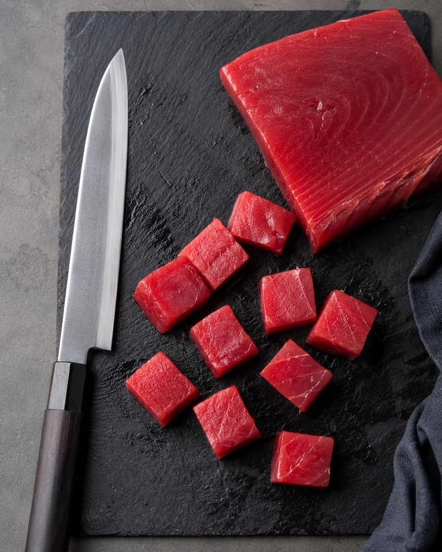 A sharp knife and cubes of raw tuna are arranged on a black slate board, with a larger block of tuna and a dark cloth nearby.