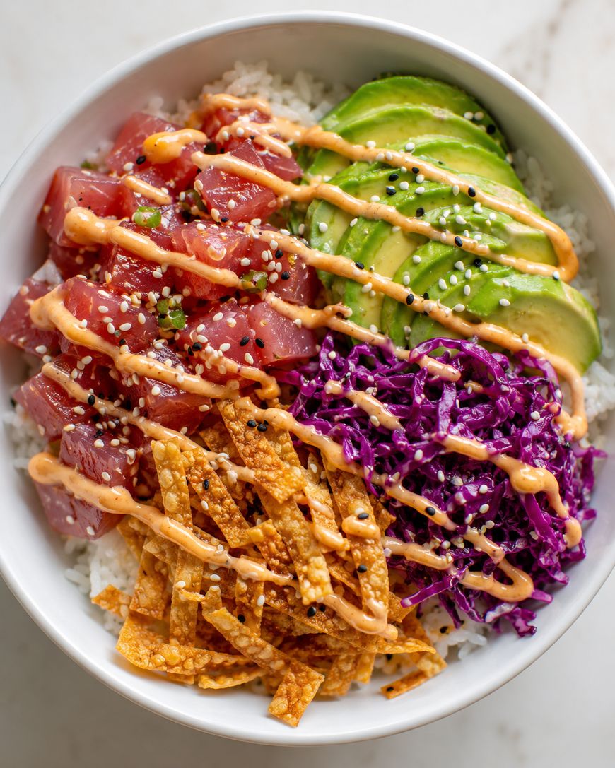 A poke bowl with cubed raw tuna, sliced avocado, shredded purple cabbage, crispy wonton strips, rice, and a drizzle of creamy orange sauce, garnished with black and white sesame seeds.