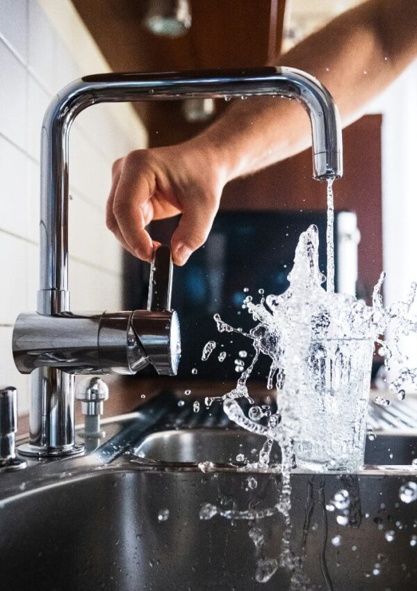 A hand turns on a kitchen faucet, causing water to splash dramatically as it pours into a glass sitting in a stainless steel sink.
