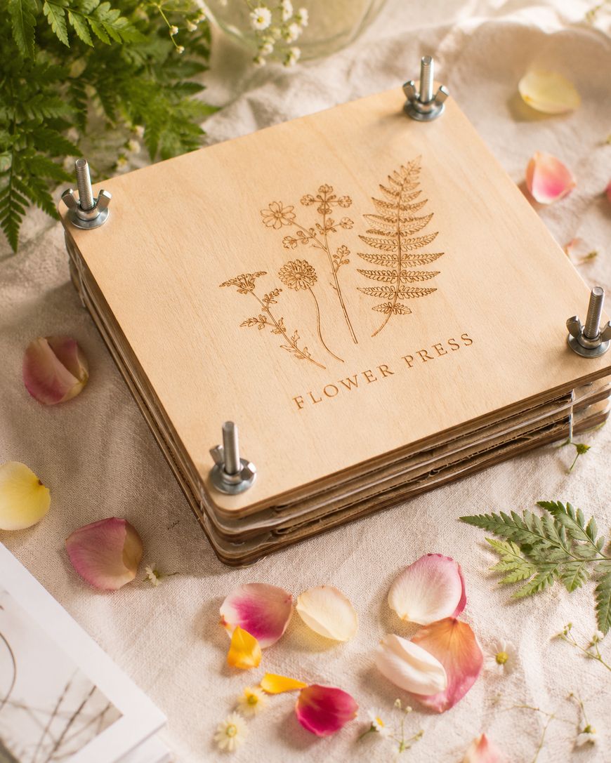 A wooden flower press with engraved botanical designs and Flower Press text sits on a fabric surface, surrounded by scattered flower petals, leaves, and a small bunch of ferns.