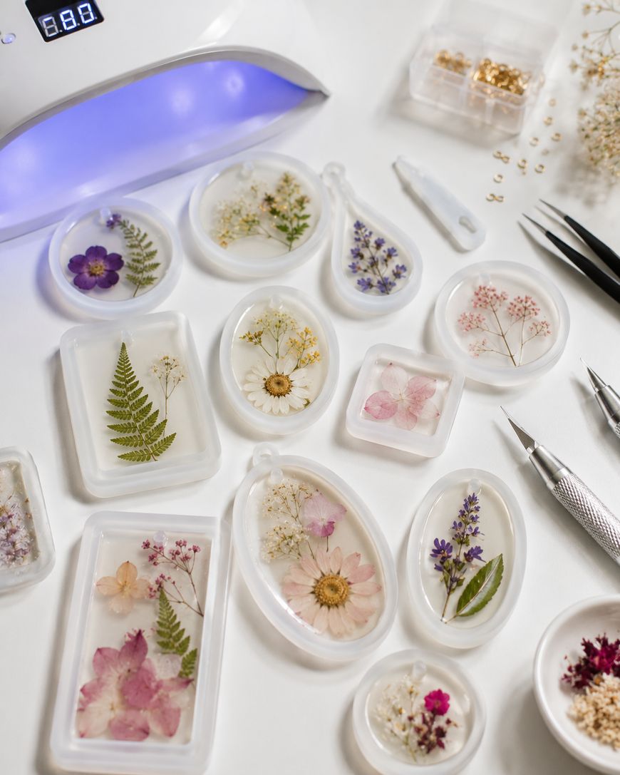 Various resin pendants with pressed flowers and leaves are arranged on a white surface alongside crafting tools, tweezers, and a UV lamp. Small containers with gold findings and tiny dried flowers are also visible.