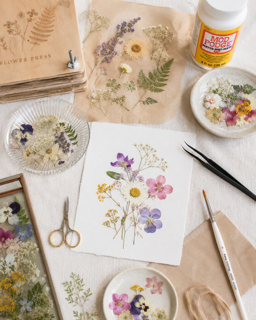 A flat lay of dried and pressed flowers, scissors, tweezers, brushes, Mod Podge glue, and paper showing a floral arrangement. Art supplies and flower petals are neatly arranged on a light surface.
