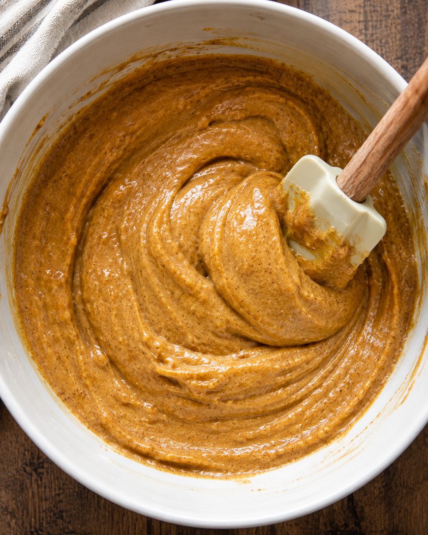 A white mixing bowl filled with thick, spiced pumpkin batter being stirred with a rubber spatula, on a wooden surface.