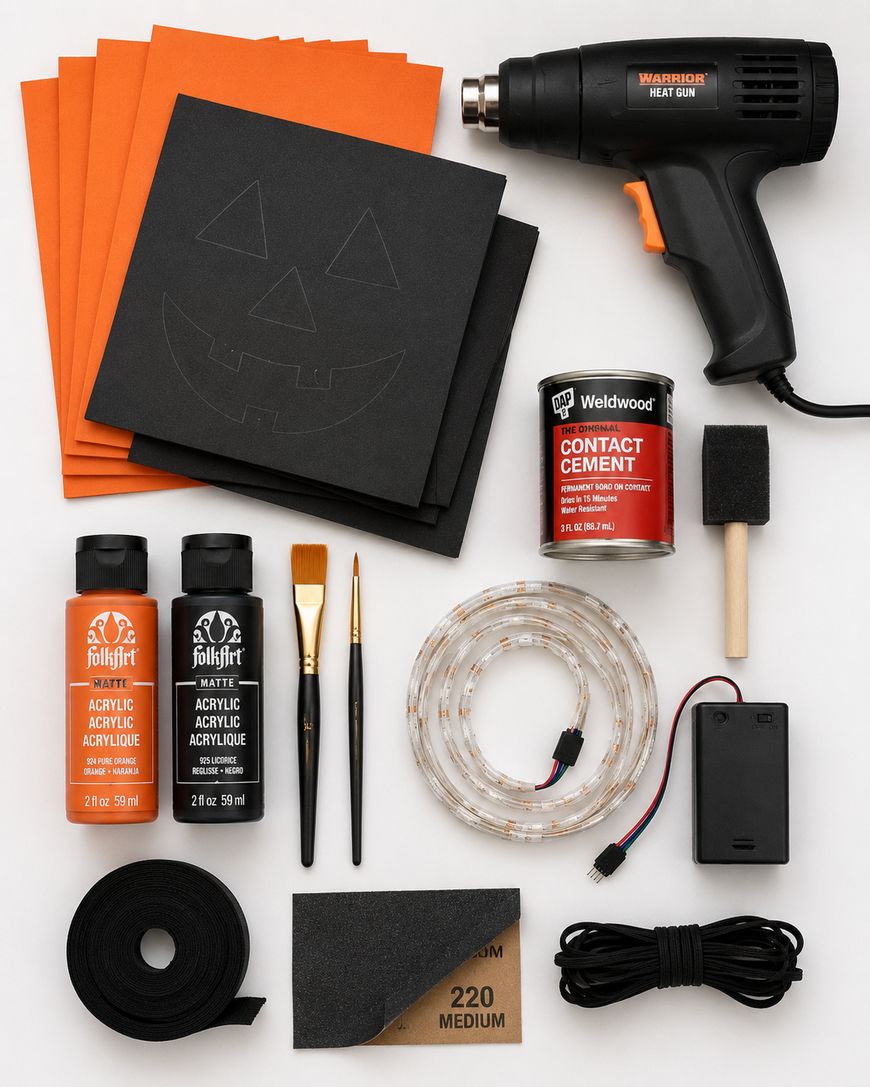 Flat lay of Halloween craft supplies: orange and black paper, heat gun, glue, acrylic paints, paintbrushes, foam brush, LED strip, battery pack, sandpaper, black tape, and contact cement, arranged neatly on a white background.