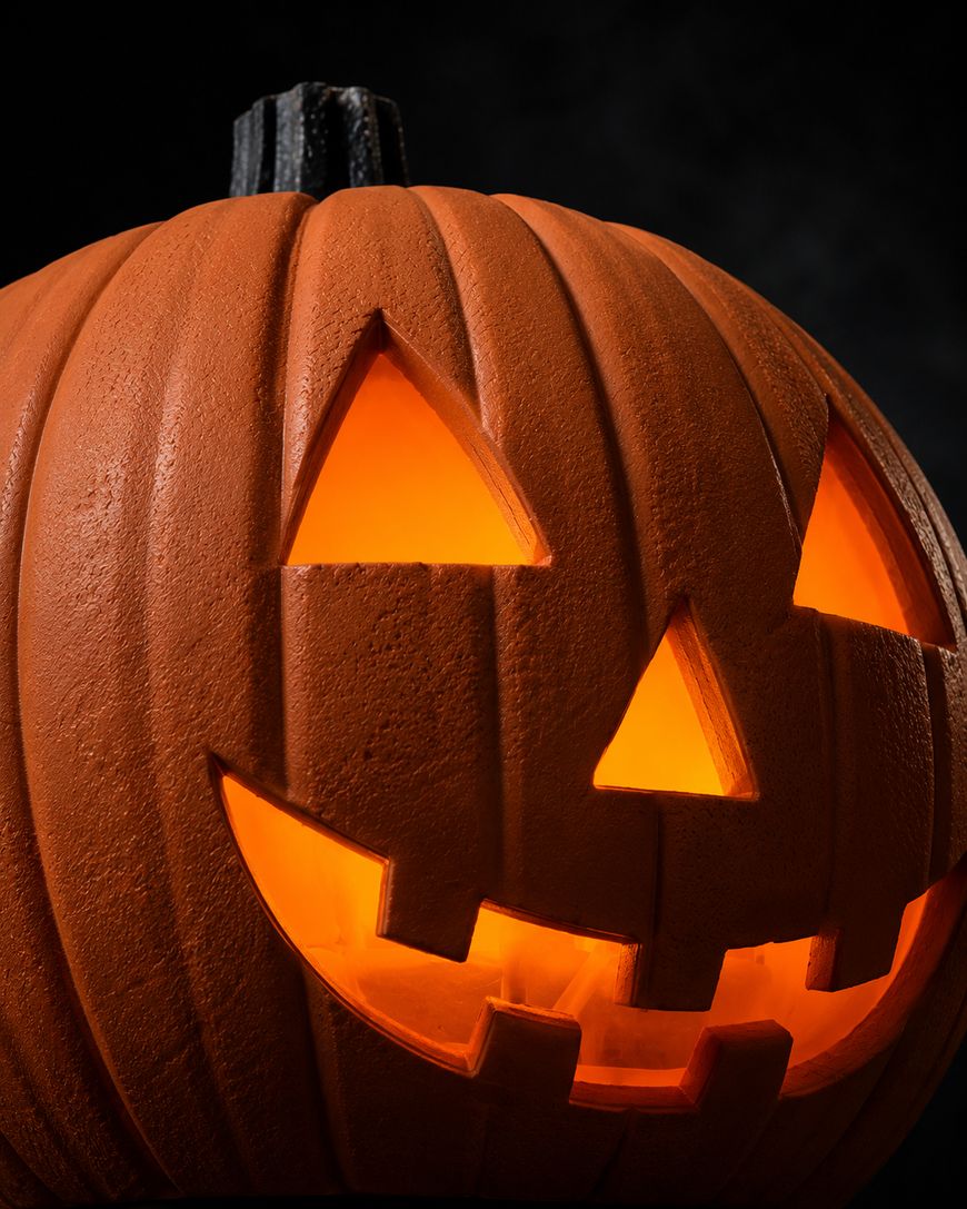 A close-up of a carved Halloween pumpkin, or jack-o-lantern, with a glowing light inside. The pumpkin has triangular eyes and nose, and a smiling mouth with square teeth. The background is dark.