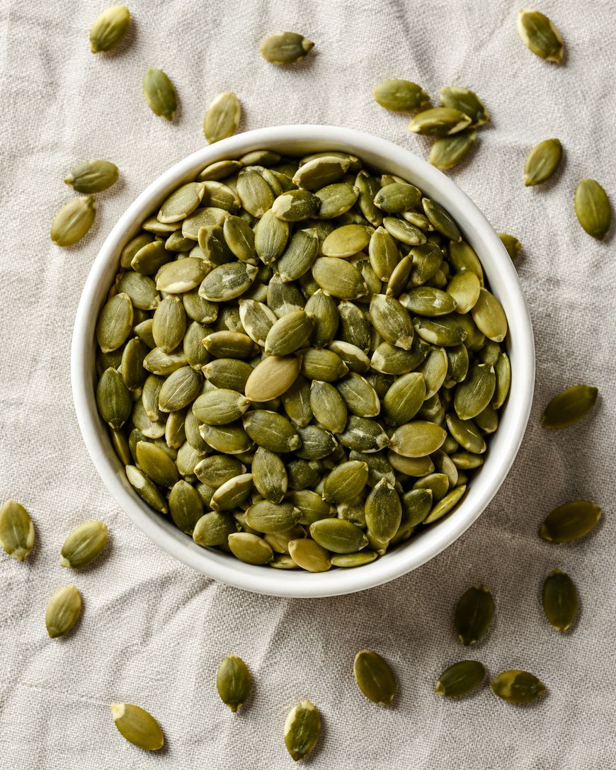 A white bowl filled with green pumpkin seeds sits on a light-colored fabric, with several seeds scattered around the bowl.