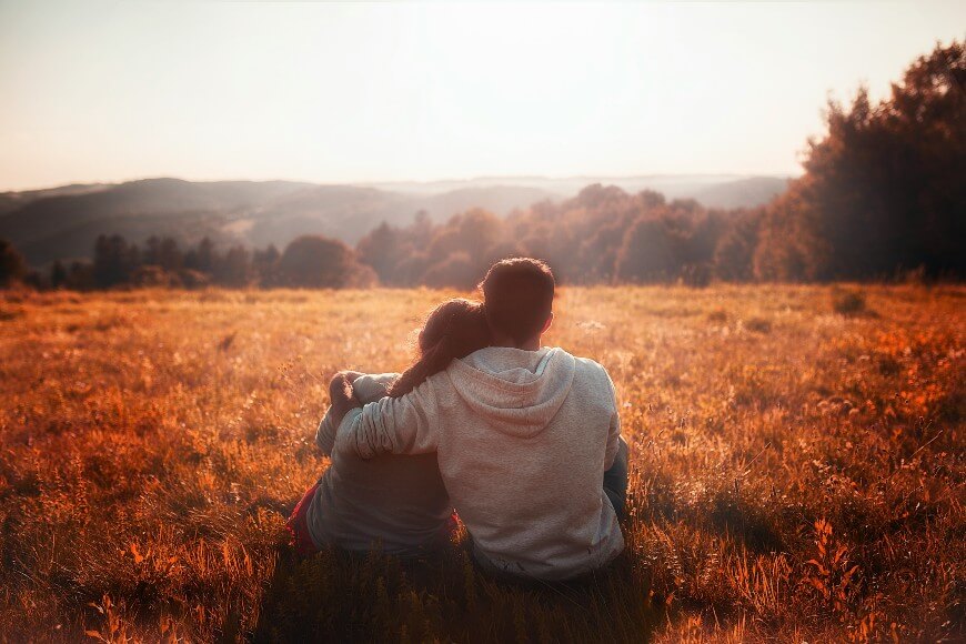 Two people sit close together in a grassy field at sunset, with one person’s arm around the other. They face away, looking out at distant hills and trees under a warm, golden sky.