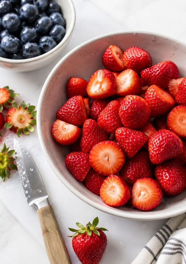 A bowl filled with sliced strawberries sits next to a bowl of blueberries, perfect for a Red White and Blue Trifle. A knife and strawberry tops rest on a cutting board, with a whole strawberry and striped cloth nearby on a white surface.