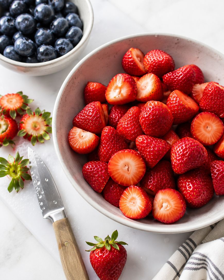 A bowl of halved strawberries next to a bowl of blueberries. A knife and strawberry tops are on the side, resting on a white surface with a striped cloth nearby.