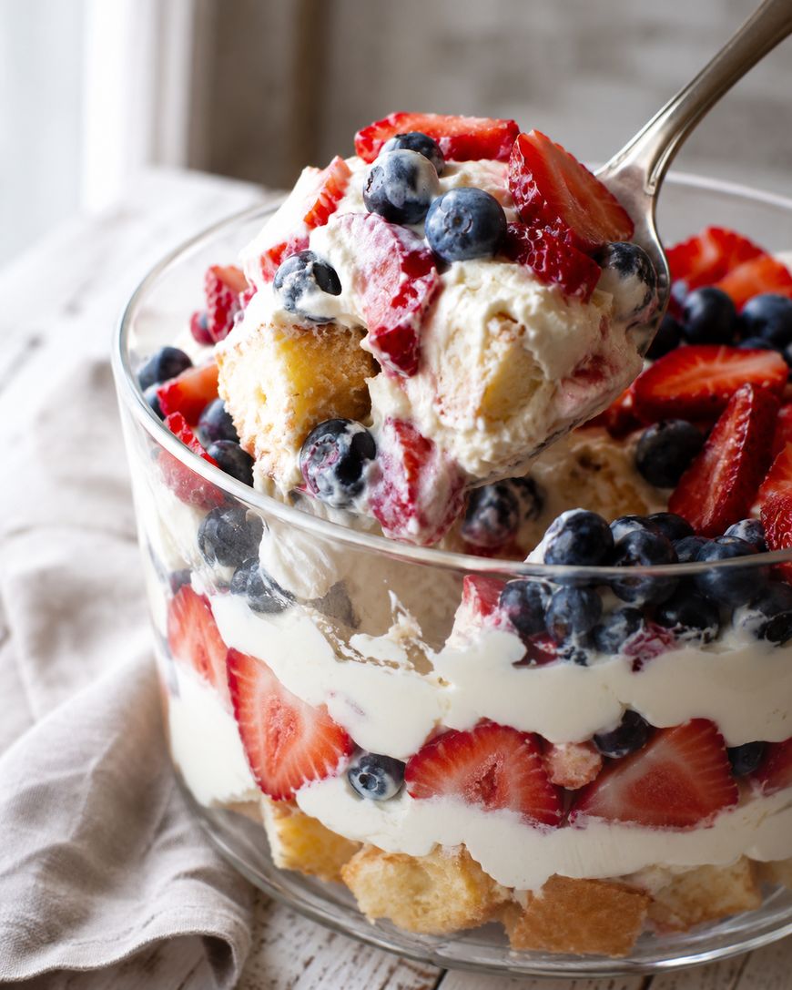 A glass bowl filled with strawberry and blueberry trifle, featuring layers of sliced strawberries, blueberries, whipped cream, and cake pieces. A spoon is scooping out a portion.