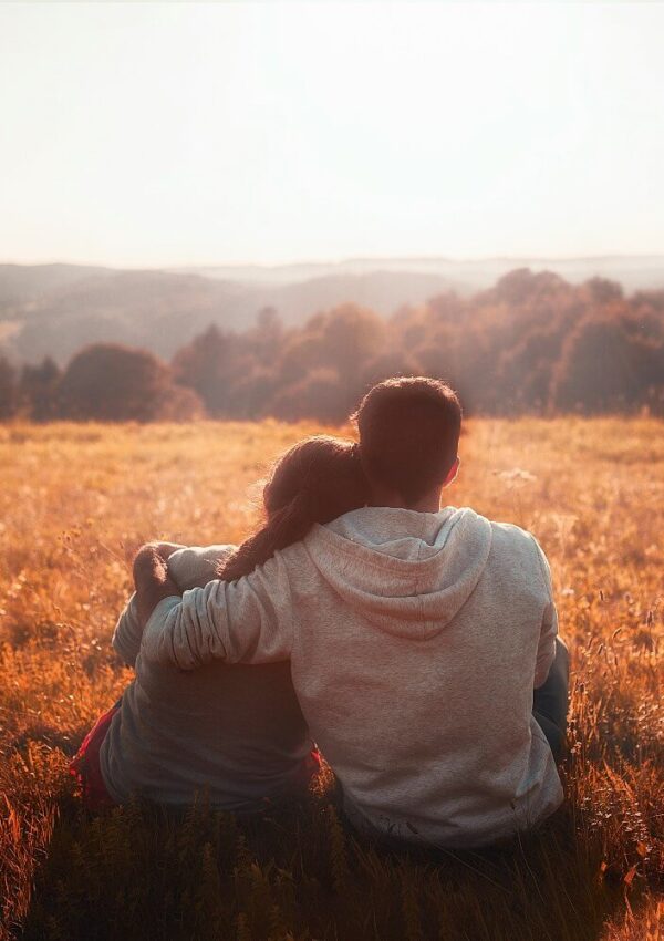 A couple sits in a grassy field at sunset, with the man’s arm around the woman. They are facing away, looking toward distant hills and a softly lit sky. The scene feels peaceful and intimate.