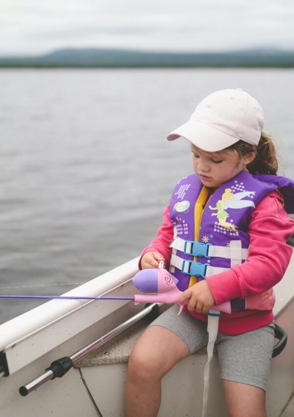 A young child wearing a life jacket, pink hoodie, and cap sits in a boat while holding a small purple fishing rod over the side, focused on fishing on a calm lake.