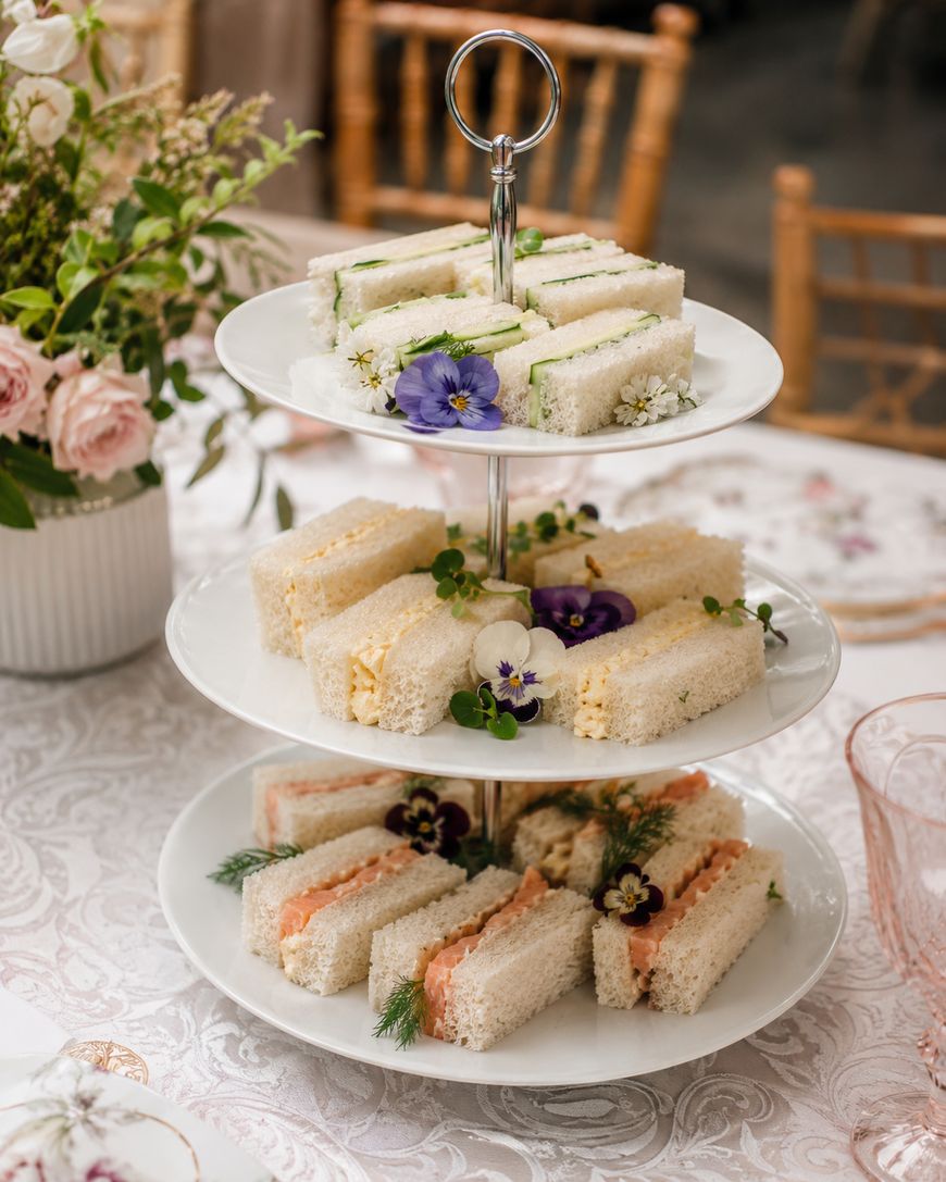 A three-tiered tray holds assorted finger sandwiches garnished with edible flowers and herbs, set on a lace tablecloth beside a floral arrangement and wooden chairs.