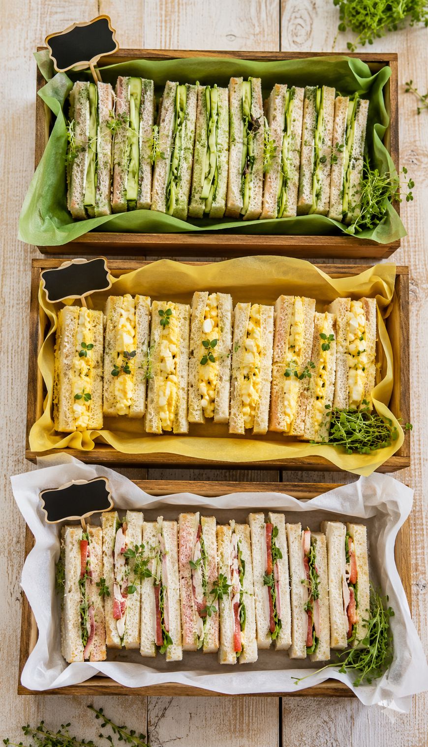 Three wooden trays of assorted tea sandwiches. The top tray has sandwiches with green filling, the middle has egg salad sandwiches, and the bottom has sandwiches with various meats and vegetables. Each tray is garnished with fresh greens.