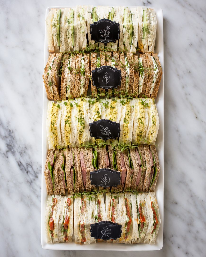 A rectangular platter with rows of assorted tea sandwiches, made with white and brown bread, filled with various vegetables and greens, and labeled with small black signs. Marble surface background.