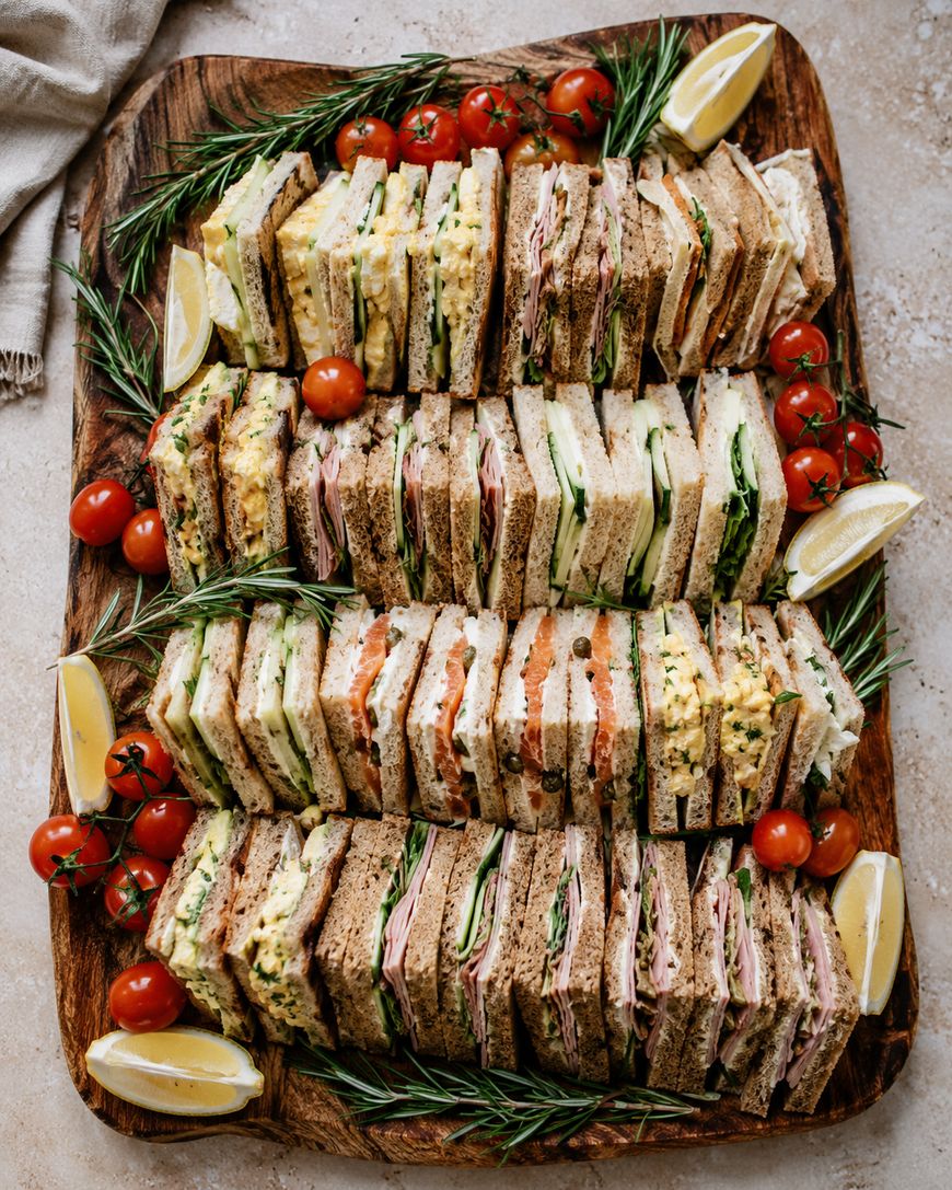 A wooden platter filled with assorted sandwich halves, garnished with fresh rosemary, lemon wedges, and cherry tomatoes. The sandwiches contain various fillings, including greens, eggs, ham, and tomatoes.