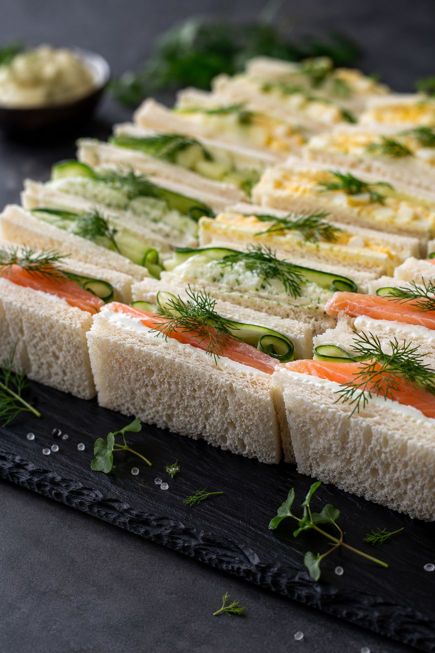 Assorted finger sandwiches on a slate tray, including ones with salmon and dill, cucumber slices, and egg salad, all made with white bread and garnished with fresh herbs.