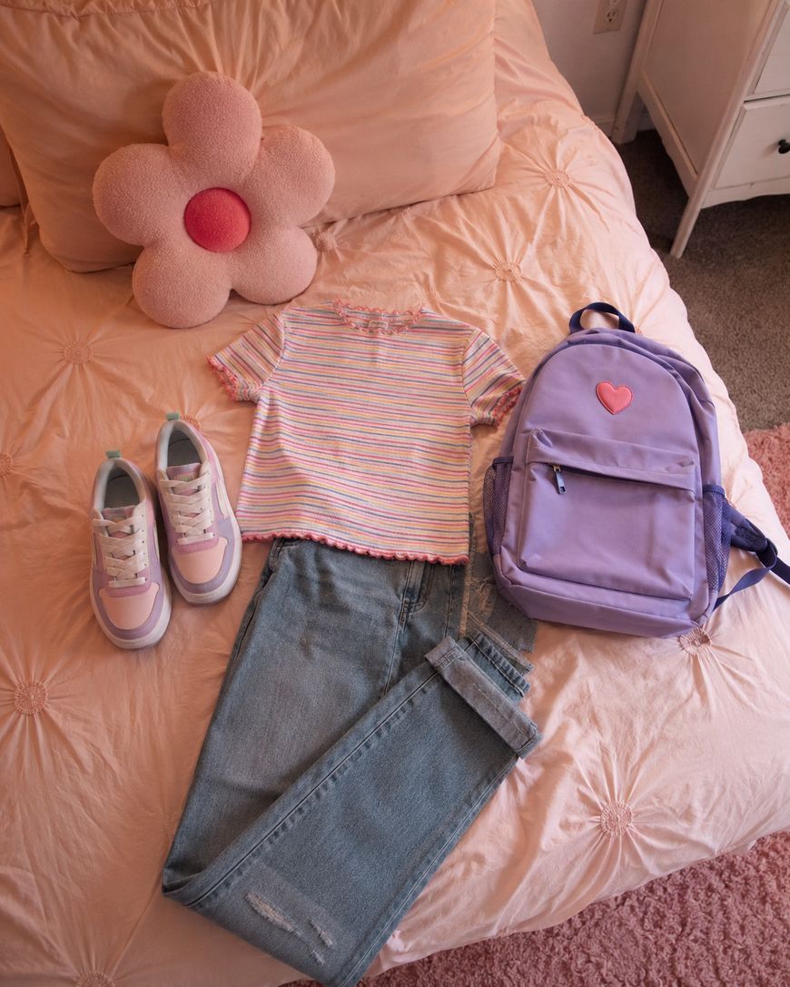 A pastel-themed outfit lies on a pink bedspread: striped t-shirt, light blue jeans, purple sneakers, and a lavender backpack with a heart patch. A flower-shaped pillow sits at the top of the bed.