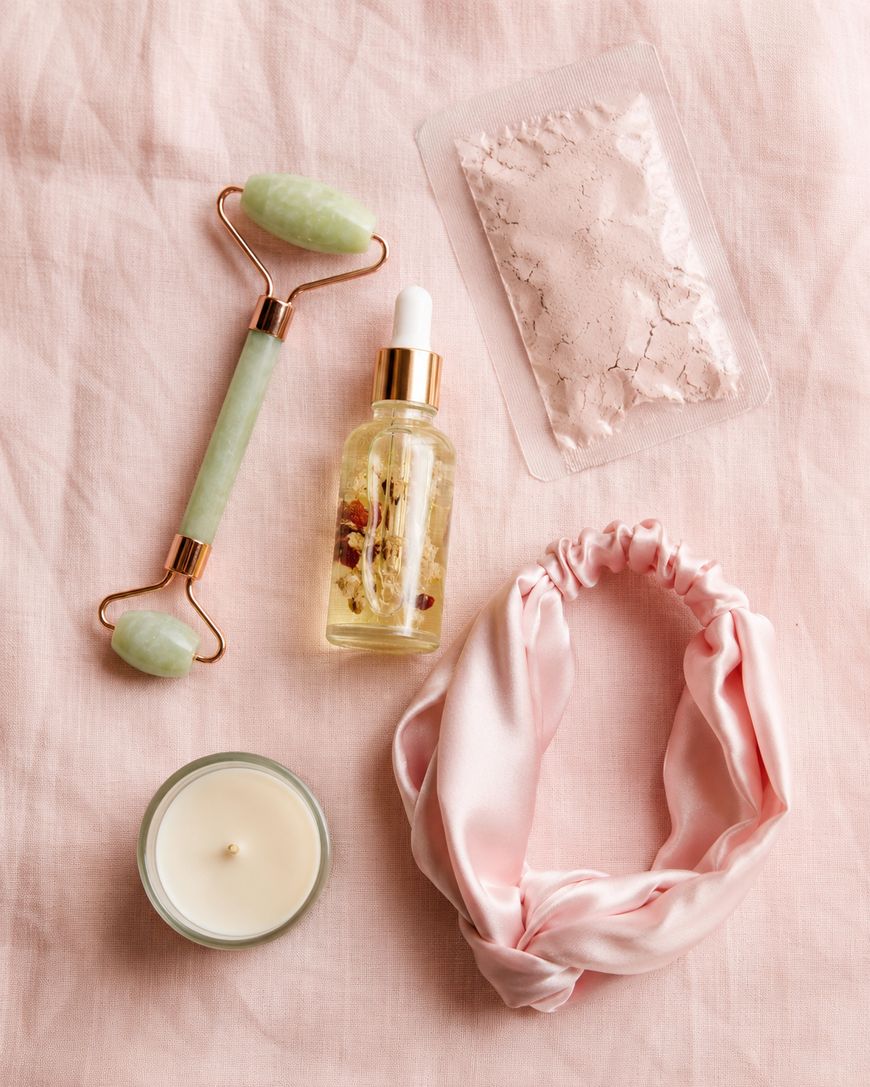 A jade facial roller, a dropper bottle with oil and herbs, a packet of powder, a lit candle, and a pink satin headband arranged on a soft pink fabric background.