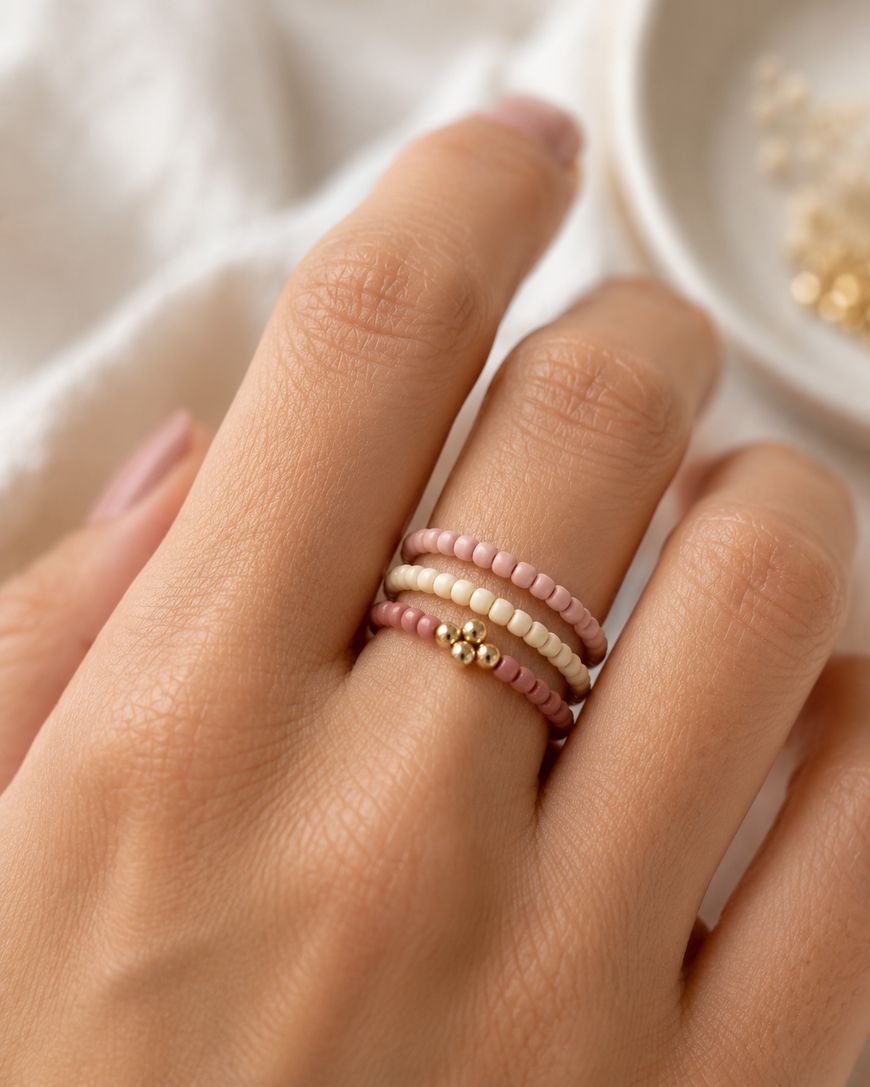 A close-up of a hand wearing three beaded rings in pink, cream, and gold tones. The rings are stacked on the ring finger, and the background is softly blurred with neutral colors.