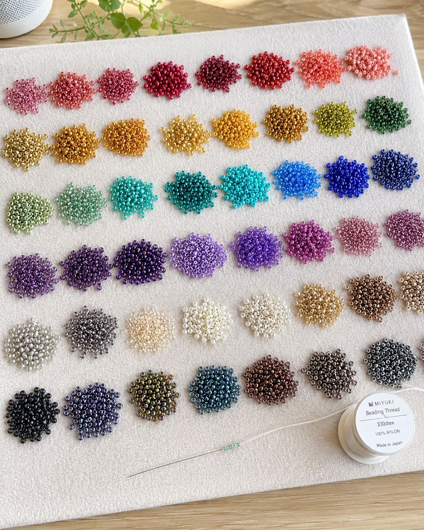 A display of colorful seed beads arranged in neat rows by shade on a white surface, with a beading needle, clear thread, and a spool of Miyuki beading thread placed at the bottom right corner.
