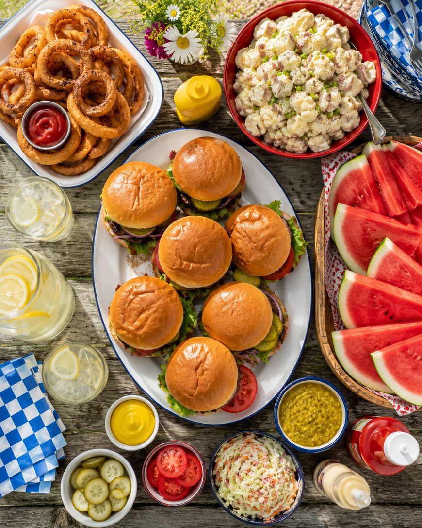 An overhead view of a picnic table with burgers, onion rings, potato salad, watermelon slices, coleslaw, sliced tomatoes, condiments, and drinks with lemon, set outdoors with flowers in the background.