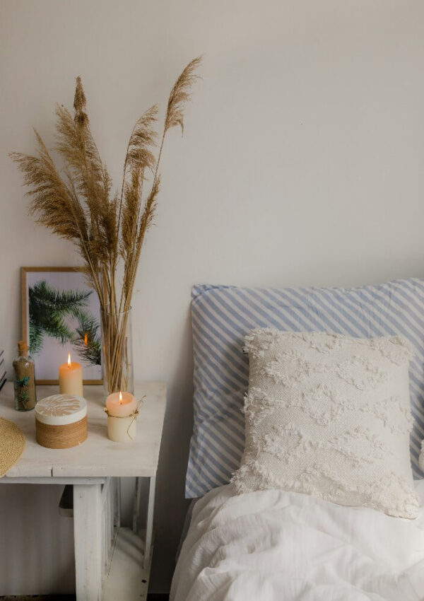A cozy bedroom scene with a white bed, textured pillows, and a side table holding lit candles, a decorative box, and a vase of dried grasses against a plain white wall.