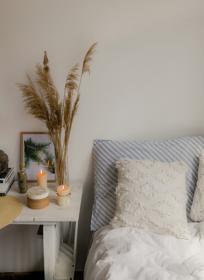 A cozy bedroom scene with a white bed, textured pillows, a white nightstand holding two lit candles, a plant in a vase, a framed pine photo, and a stack of books against a plain, light wall.