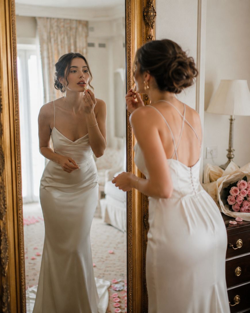 A woman in a white satin gown applies lipstick while looking into a large ornate mirror in an elegant room decorated with pink roses and soft lighting.