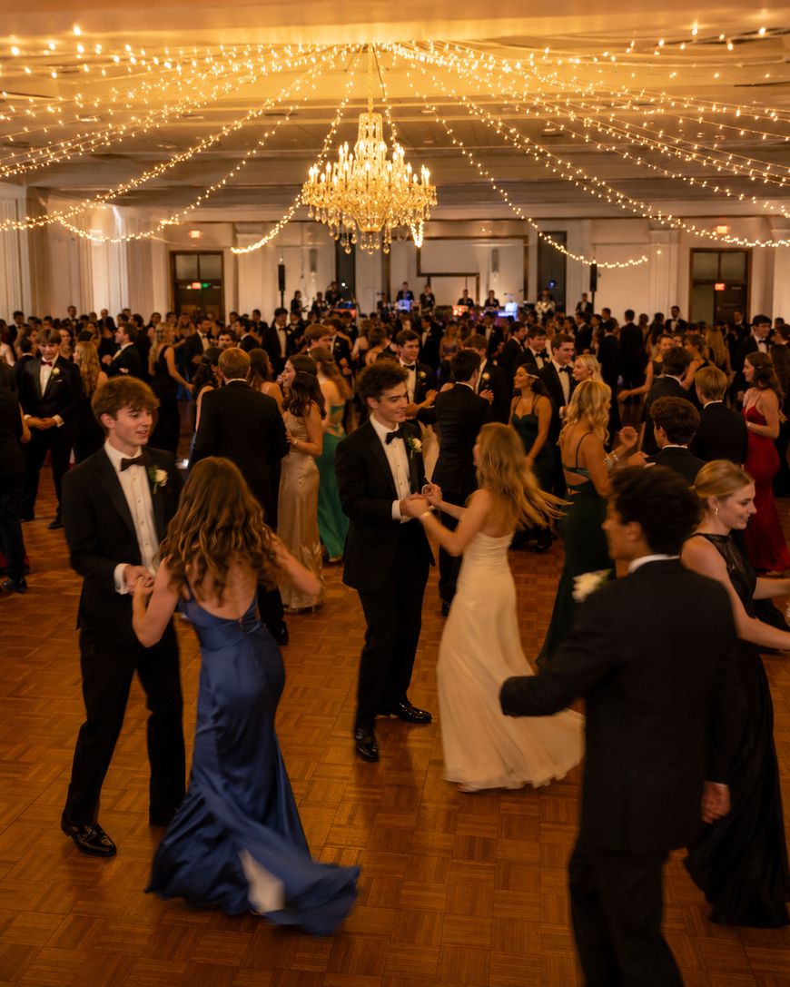 A large group of people in formal attire dance in an elegant ballroom with a chandelier and string lights overhead. The floor is crowded, and guests appear to be enjoying a lively evening event.
