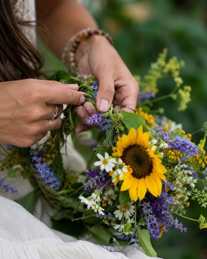 A person wearing rings and a beaded bracelet weaves a colorful flower crown with sunflowers, daisies, and wildflowers, using their hands against a green, outdoor background.
