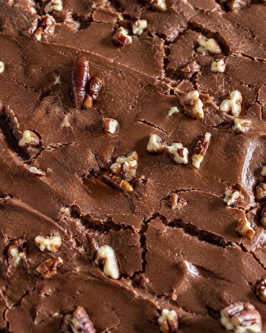 Close-up of rich, creamy chocolate fudge topped with chopped pecans, showing a smooth texture with slight cracks and scattered nut pieces.