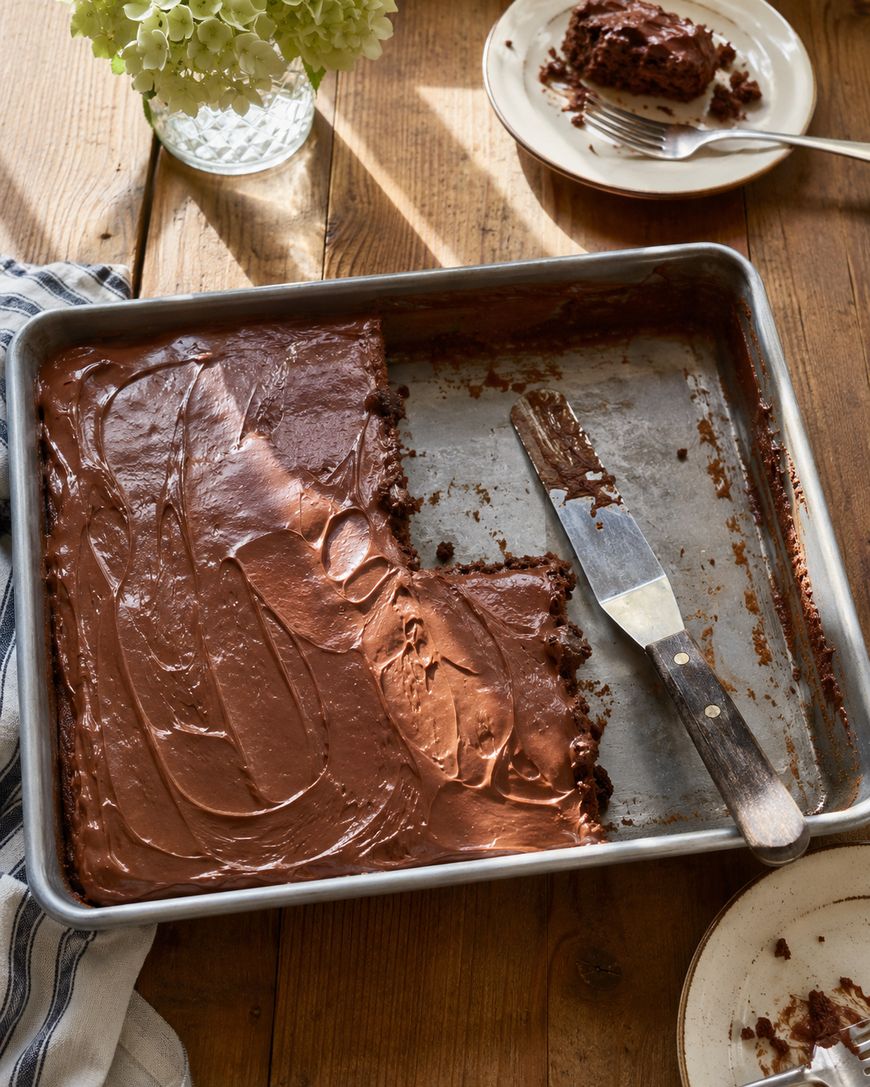 A metal baking pan with chocolate cake, partly eaten, sits on a wooden table. A metal spatula with chocolate frosting rests in the pan. Plates with cake slices and a vase of green flowers are nearby.
