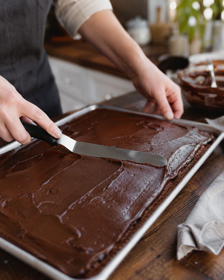A person spreads chocolate frosting on a large rectangular cake with an offset spatula in a kitchen, with a bowl of more frosting beside them.