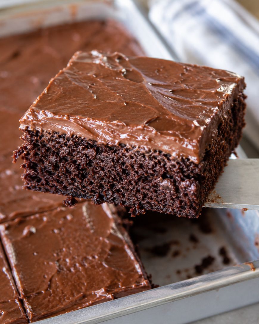 A close-up of a square piece of chocolate cake with thick chocolate frosting being lifted from a pan, showing moist, dense cake and smooth, glossy icing.