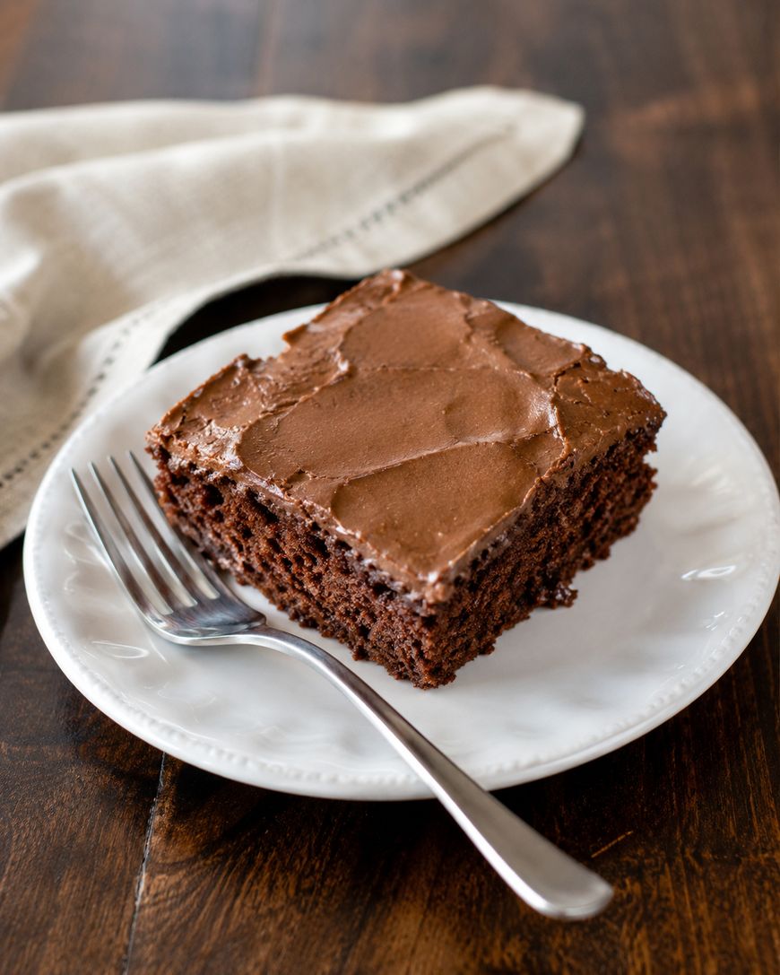 A square slice of chocolate cake with chocolate frosting sits on a white plate with a silver fork, on a dark wooden table next to a beige cloth napkin.