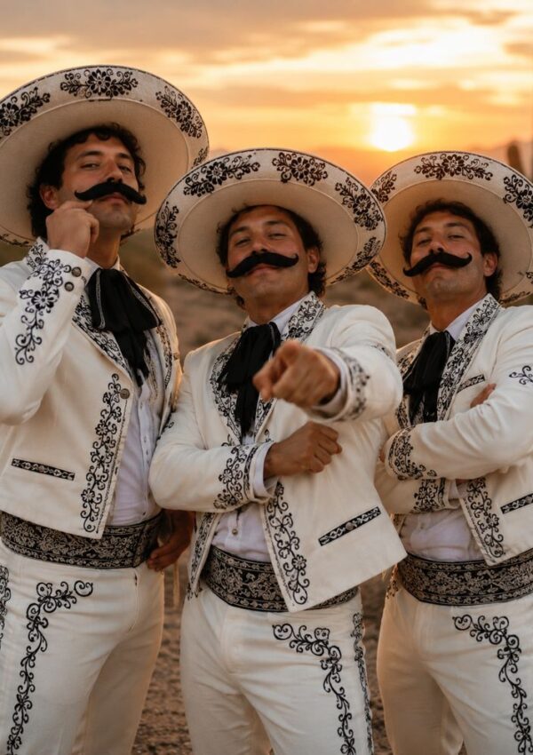 Three men in ornate white charro suits and large embroidered sombreros pose confidently outdoors at sunset—an inspiring Three People Halloween Costume with thick fake mustaches, one pointing at the camera, and cacti visible in the background.