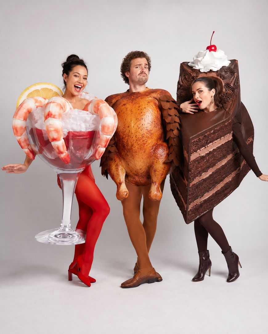 Three people in costumes: one as a shrimp cocktail with lemon, one as a roasted turkey, and one as a slice of chocolate cake with whipped cream and a cherry, all posing playfully against a plain background.
