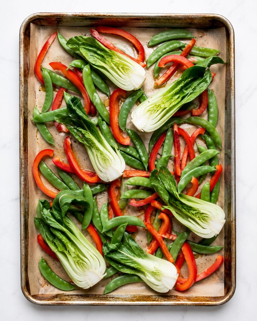 A sheet pan lined with parchment paper topped with halved baby bok choy, sliced red bell peppers, and snap peas, all arranged in a single layer, ready for roasting.