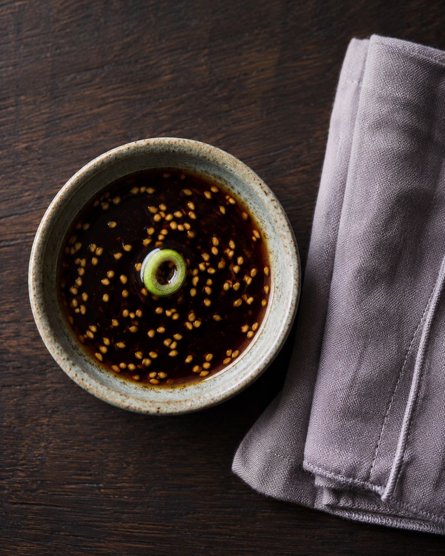 A small ceramic bowl filled with dark soy sauce, sesame seeds, and a sliced green onion, next to a neatly folded lavender cloth napkin on a dark wooden surface.
